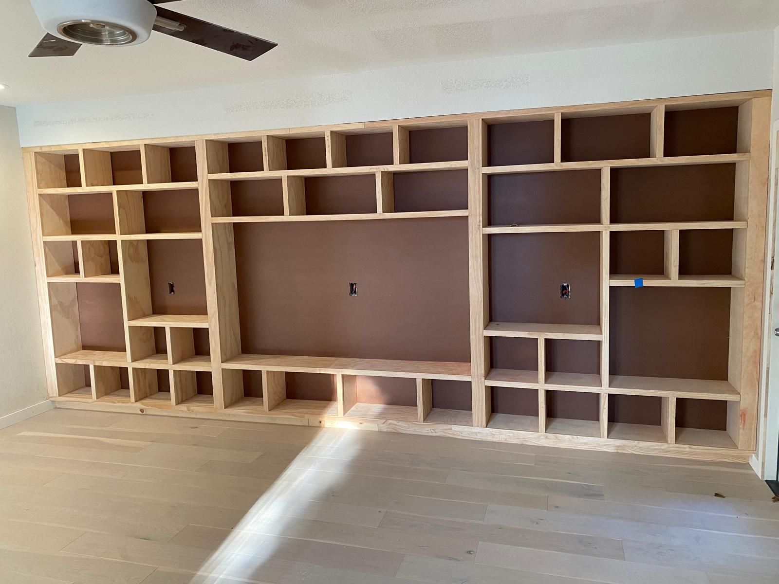 A large, unfinished wooden built-in shelving unit with a central space for a television, positioned against a white wall.