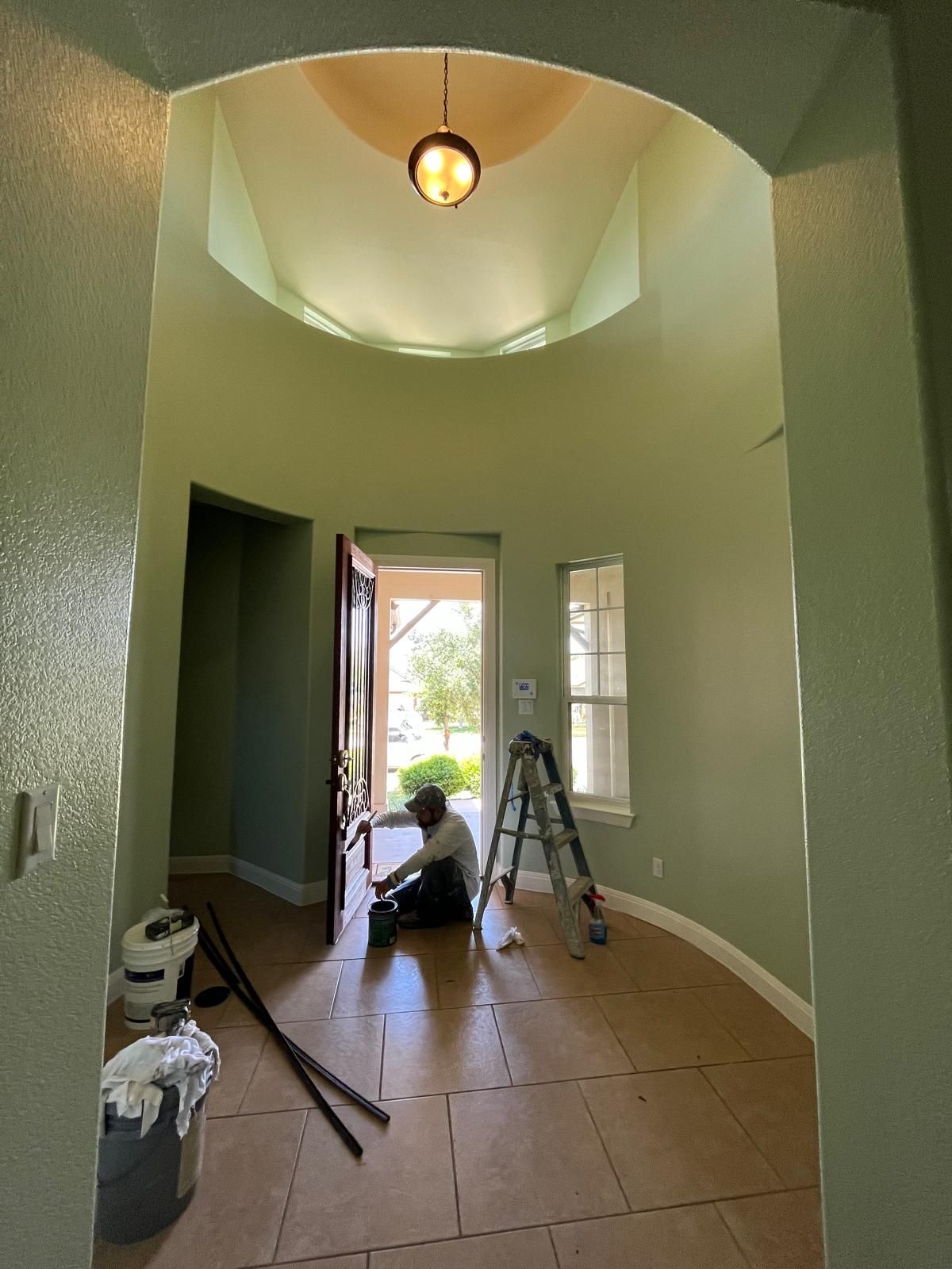 A person painting the light green walls of a high-ceilinged foyer with a ladder and supplies on the floor.