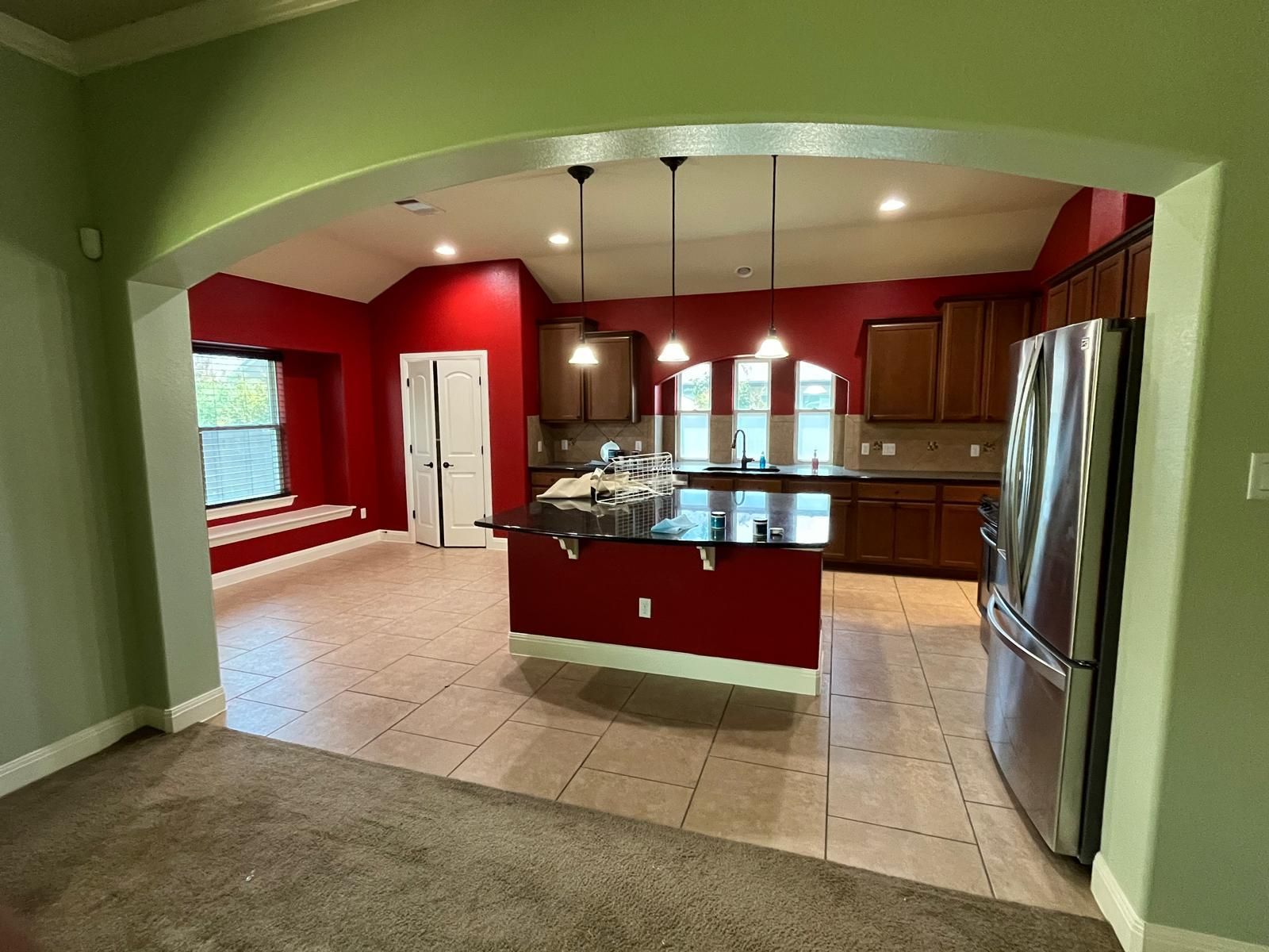 View of a red-walled kitchen with a central island and stainless steel refrigerator from a carpeted living area.