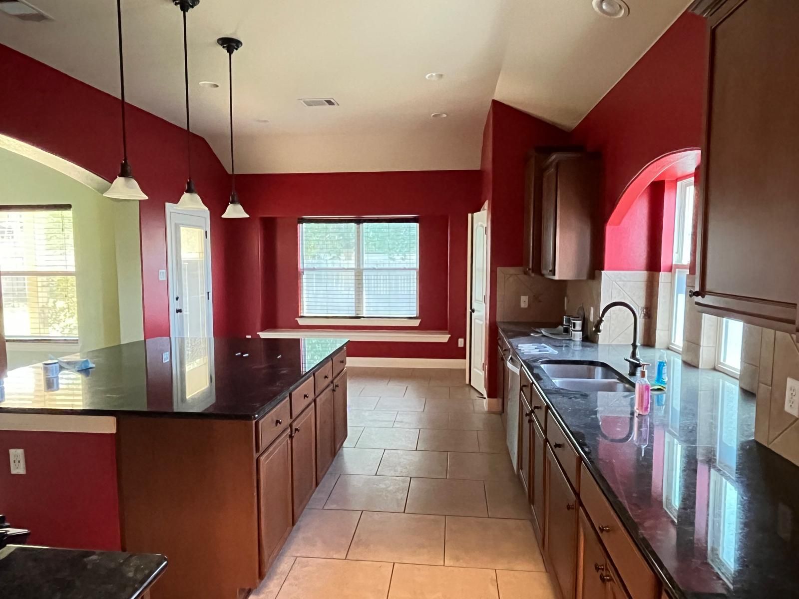 A kitchen with red walls, a dark granite island, brown cabinets, and tile floors under three pendant lights.