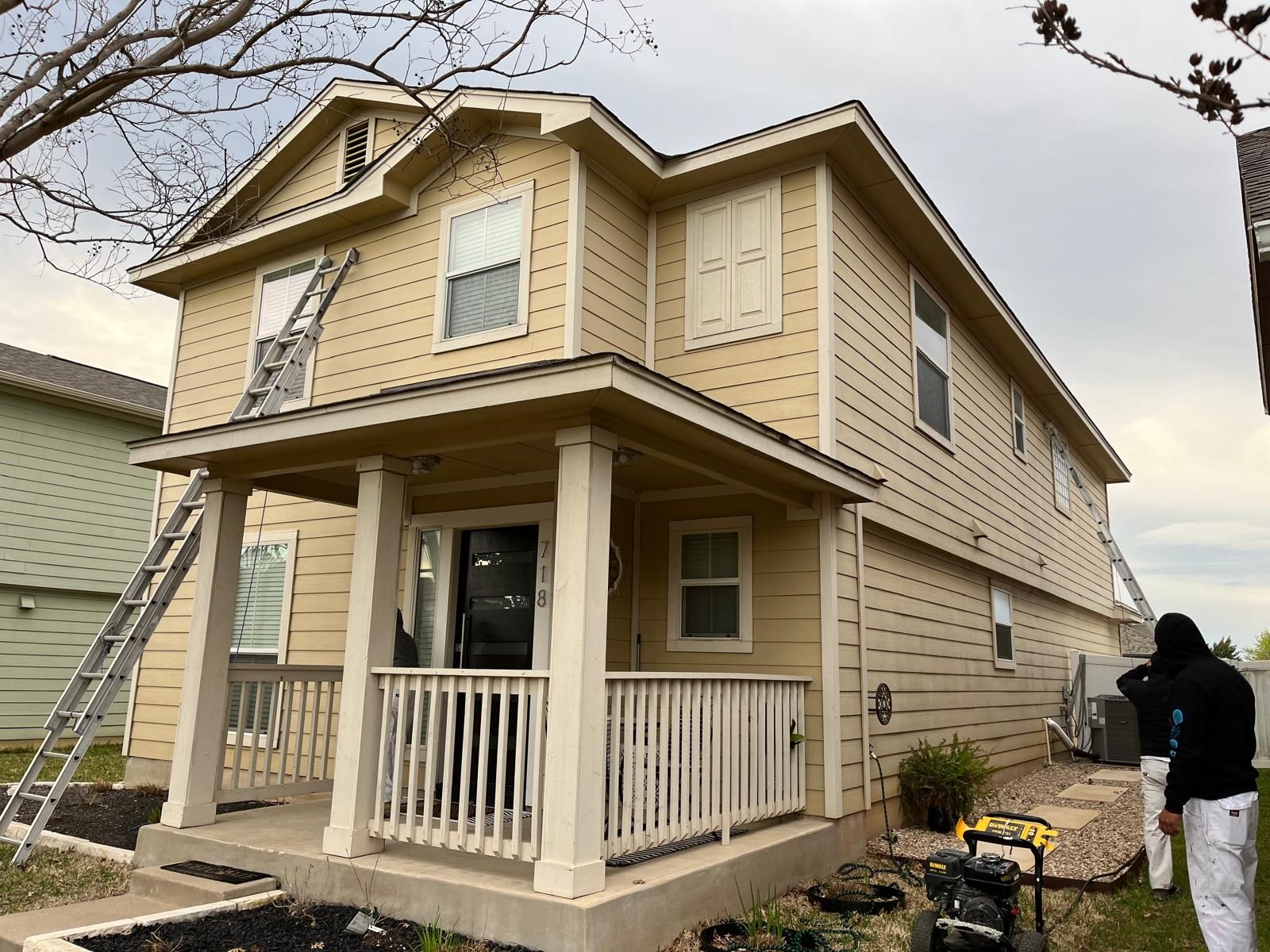 A two-story yellow house under renovation with workers, ladders, and painting equipment on the exterior.