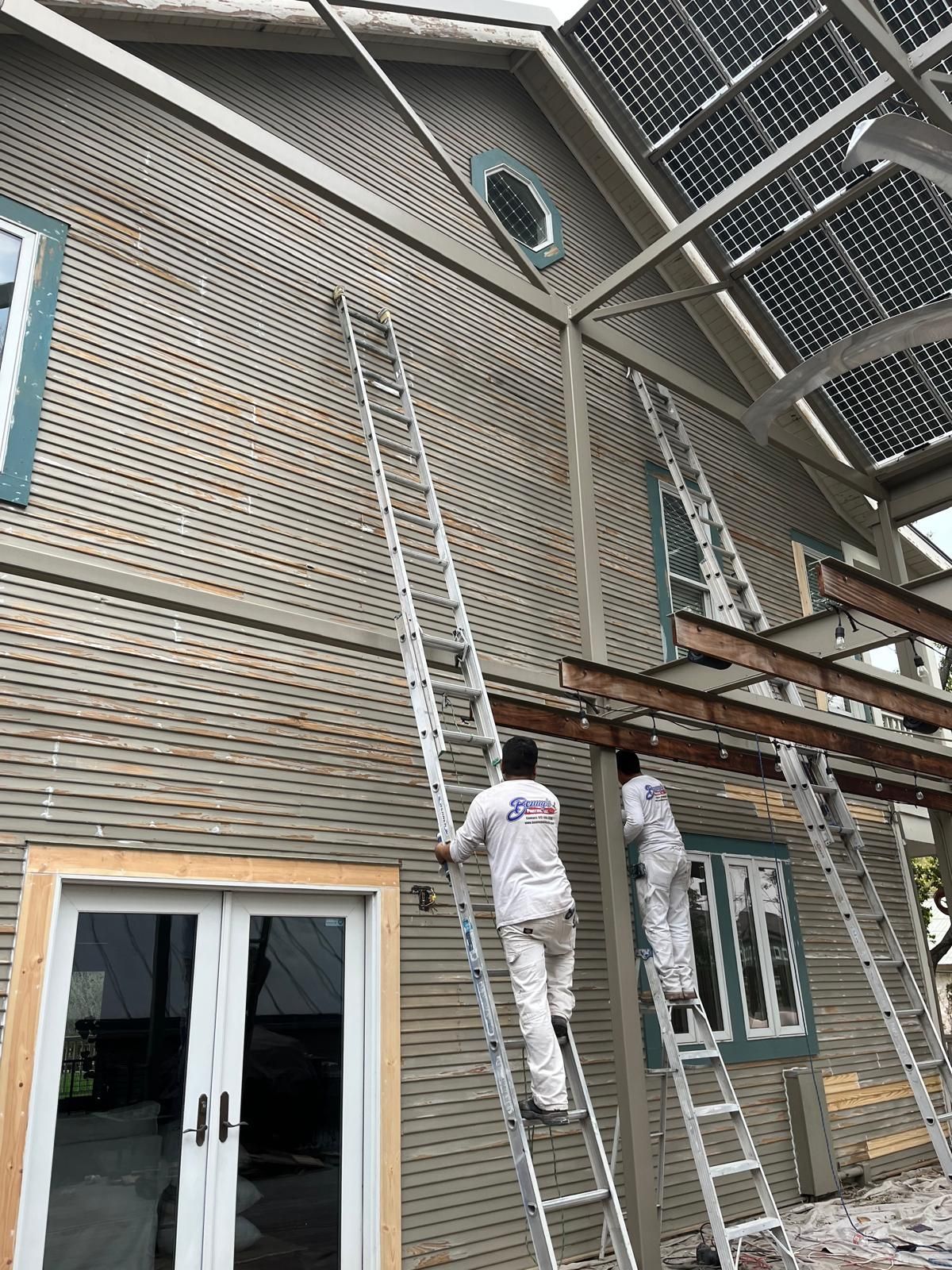 Two workers in white uniforms climb tall extension ladders against the light grey, textured exterior of a house.