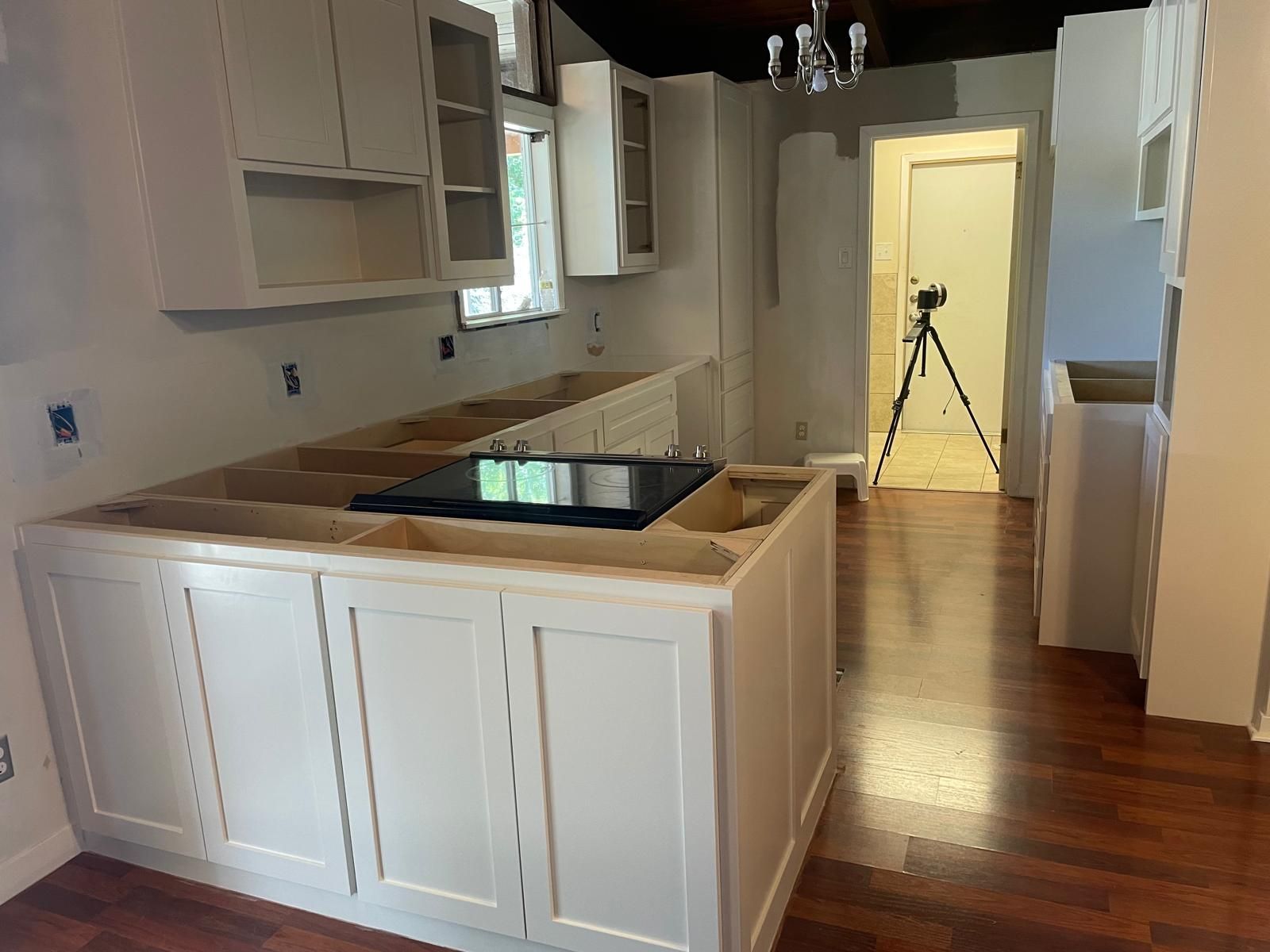 An unfinished kitchen remodel with white cabinets, a central island with a stove, and a tripod in the background.