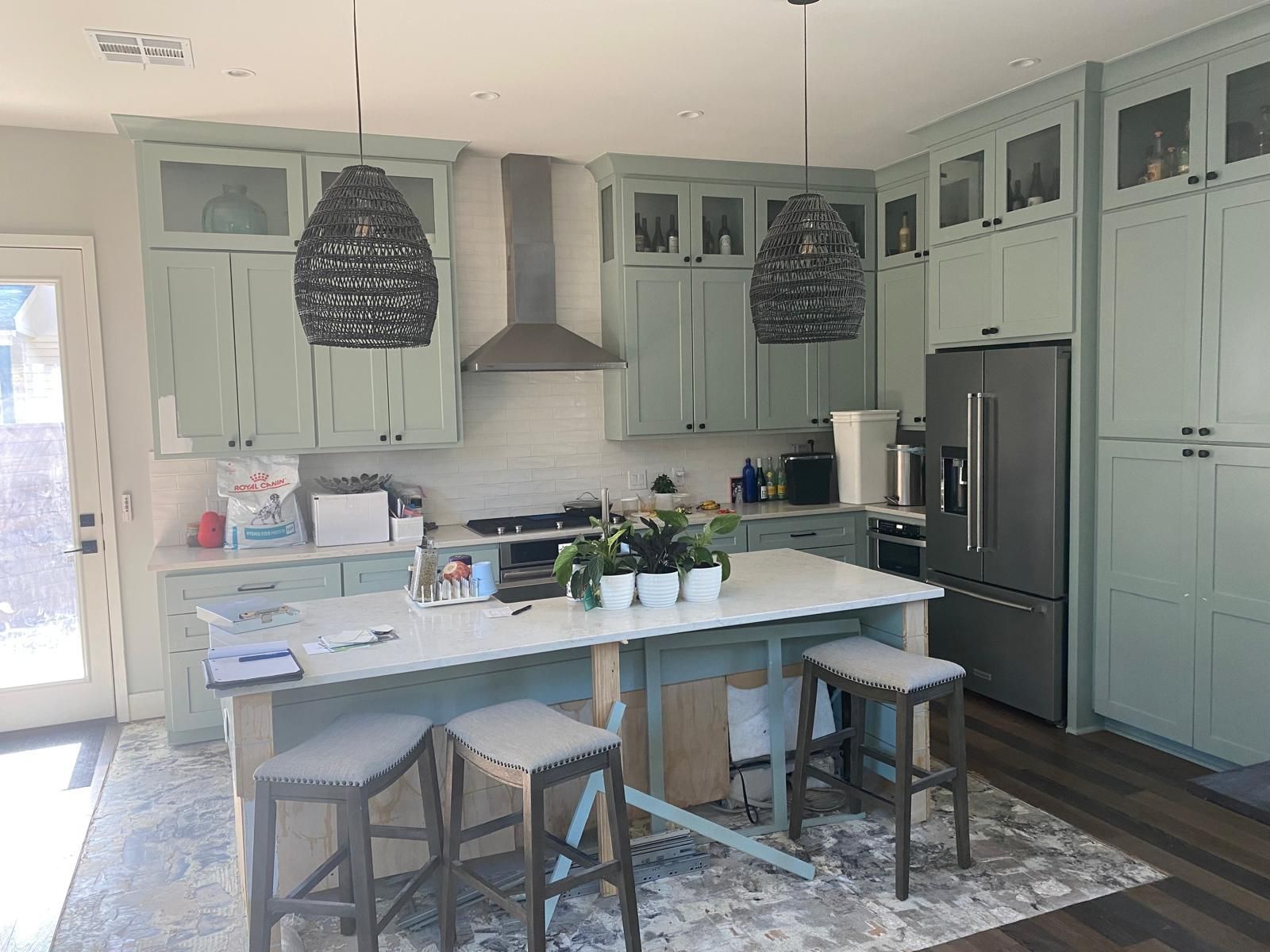 Sage green kitchen with white island, three bar stools, dark pendant lights, stainless steel appliances, and wood floors.