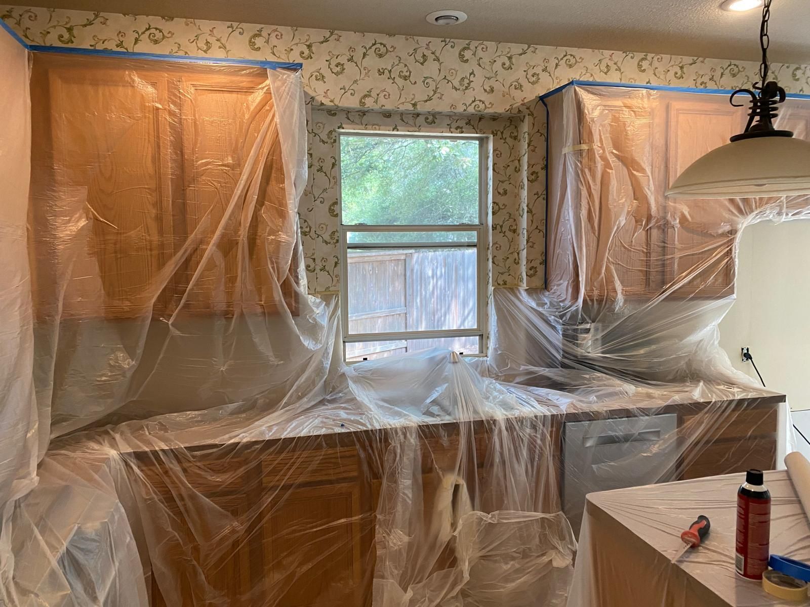 Kitchen cabinets and countertops covered in plastic sheeting for painting, with patterned wallpaper in the background.