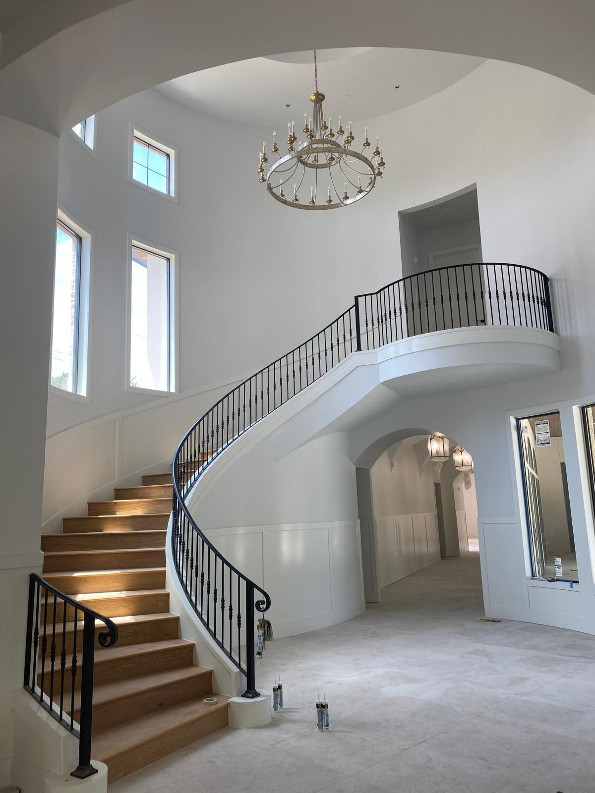 A grand curved staircase with black metal railings in a high-ceilinged foyer with white walls and a circular chandelier.
