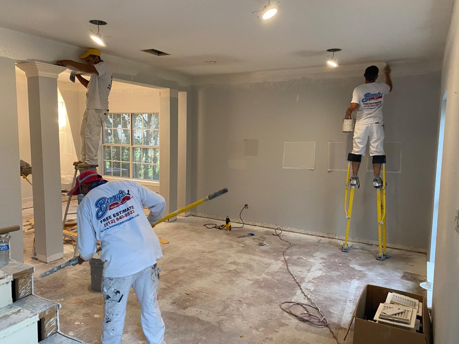 Three painters in matching white uniforms work on the walls and ceiling of a room under renovation.