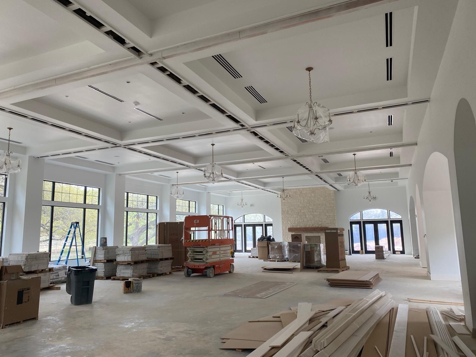 A large, unfinished interior space with white coffered ceilings, chandeliers, construction equipment, and stacked boxes.
