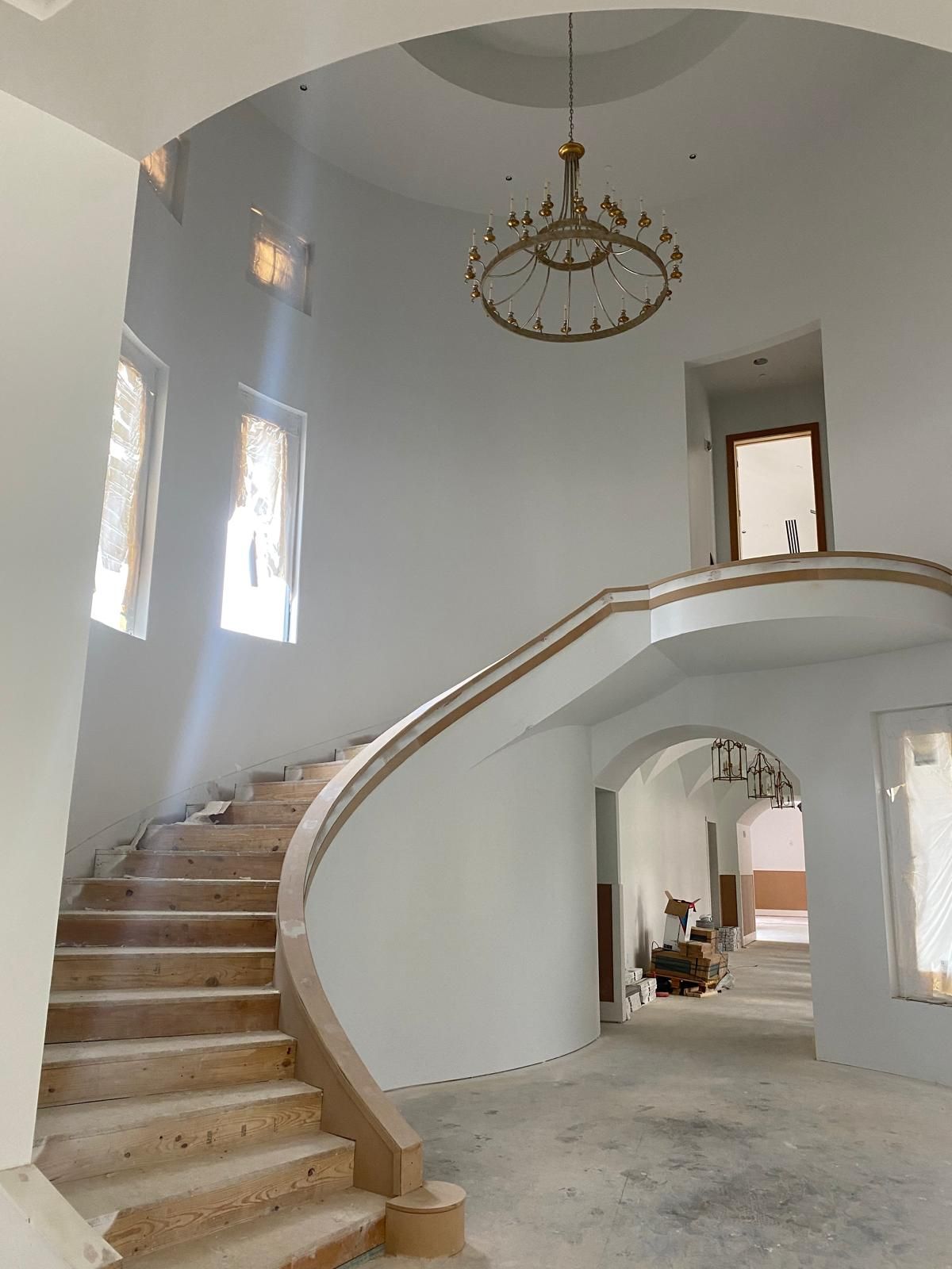 A grand, unfinished foyer with a curved staircase, tall windows, white walls, and a large circular chandelier overhead.