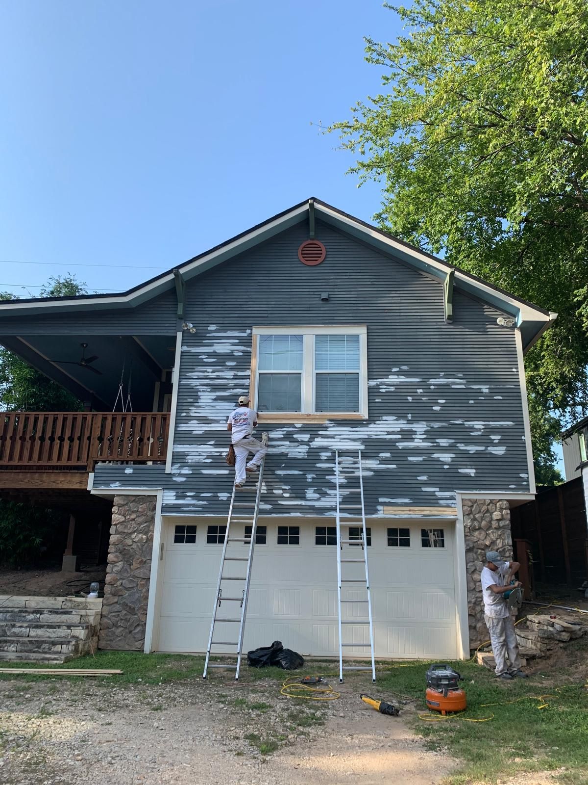 Two painters working on ladders to repaint the dark grey siding of a house above a white two-car garage.