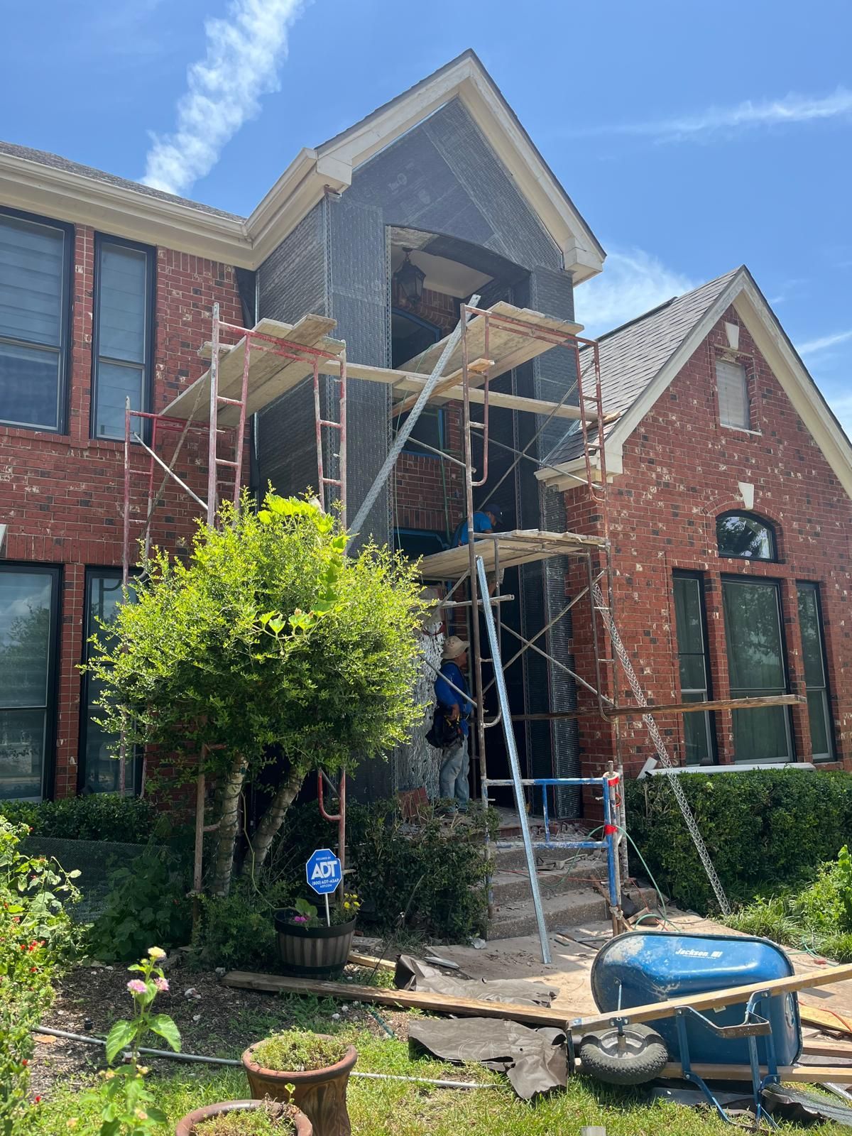 A two-story brick house with a tall, central grey-clad entrance under construction, surrounded by scaffolding and a worker.