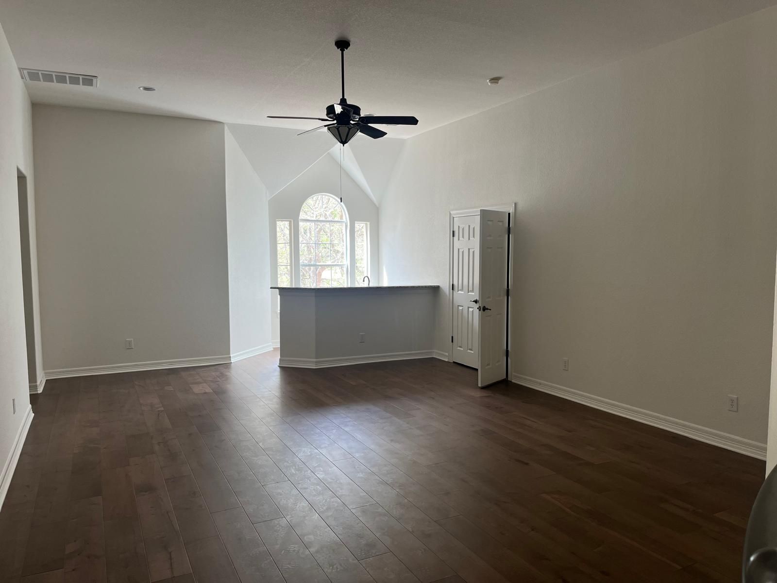 An empty, sunlit room featuring dark wood flooring, a ceiling fan, a kitchen island counter, and a white interior door.