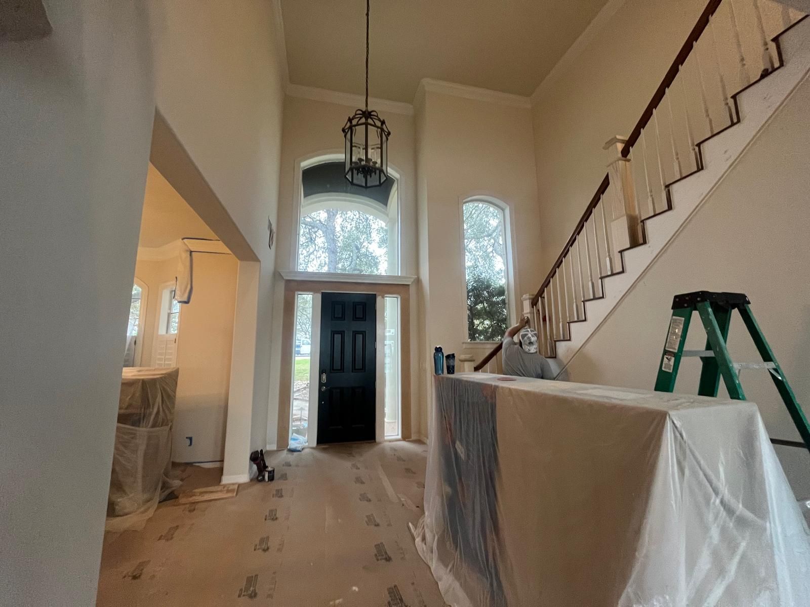 An entrance hall undergoing renovation with floor coverings, a black front door, a tall window, and a wooden staircase.
