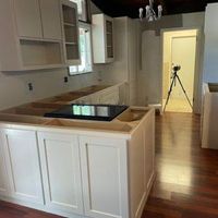 Unfinished kitchen renovation featuring white cabinets, a central island with a cooktop, and a tripod standing in a doorway.