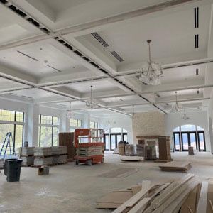 A bright, open room under construction with white coffered ceilings, chandeliers, large windows, and building materials.