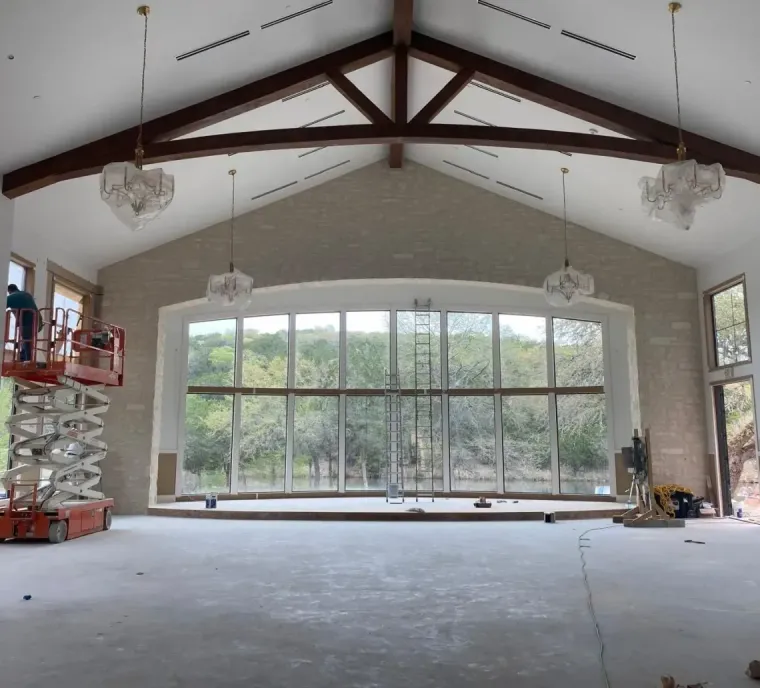An indoor construction site features a high vaulted ceiling with timber beams, a large glass window wall, and a scissor lift.
