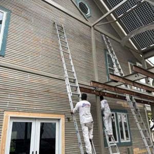 Two workers in white uniforms climb tall extension ladders against the side of a grey house to perform exterior work.