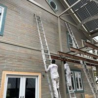 Two workers in white uniforms climb tall extension ladders against the side of a grey house to perform exterior work.