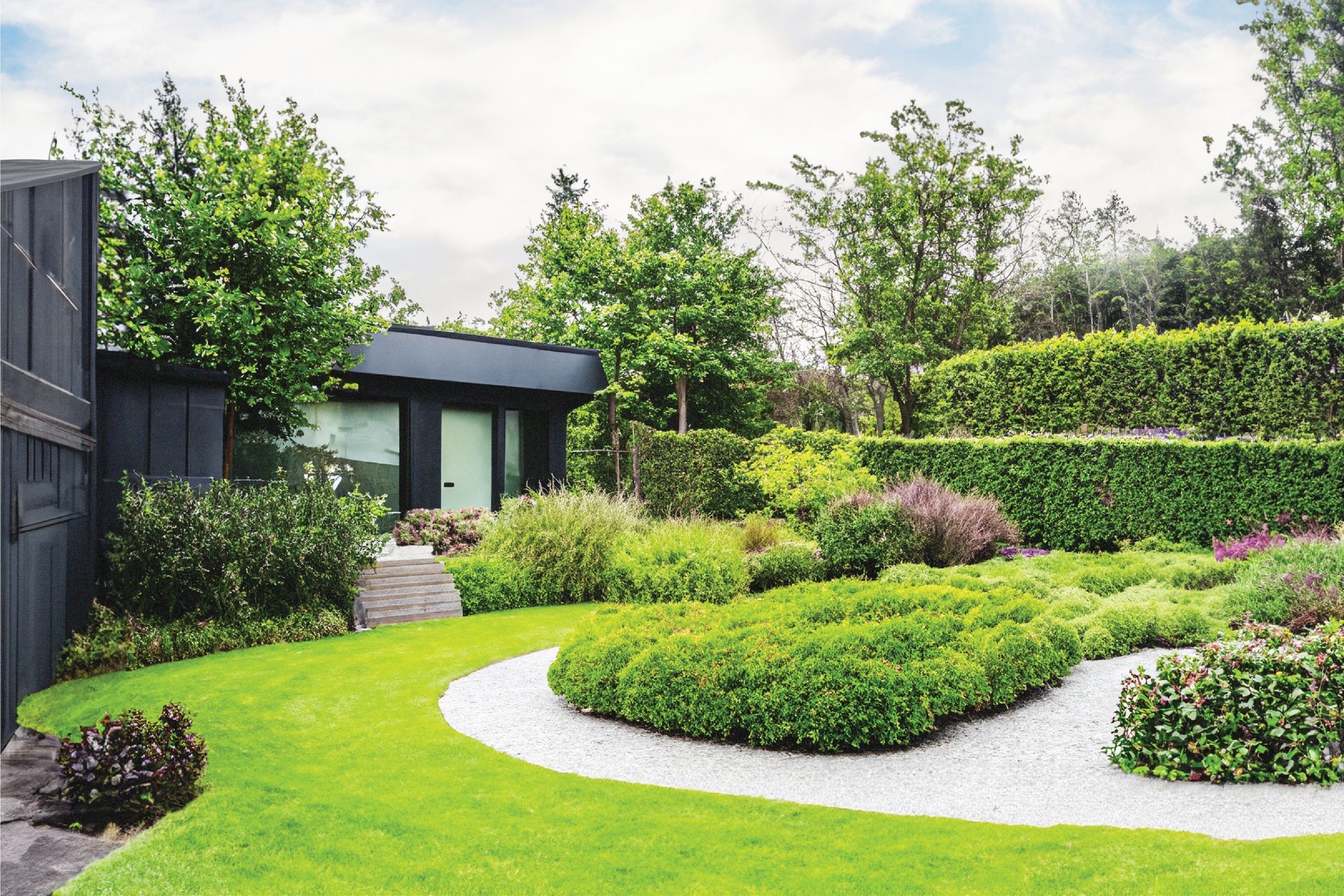Lush green garden with curving white gravel path, shrubs, trees, and a small modern house in the background