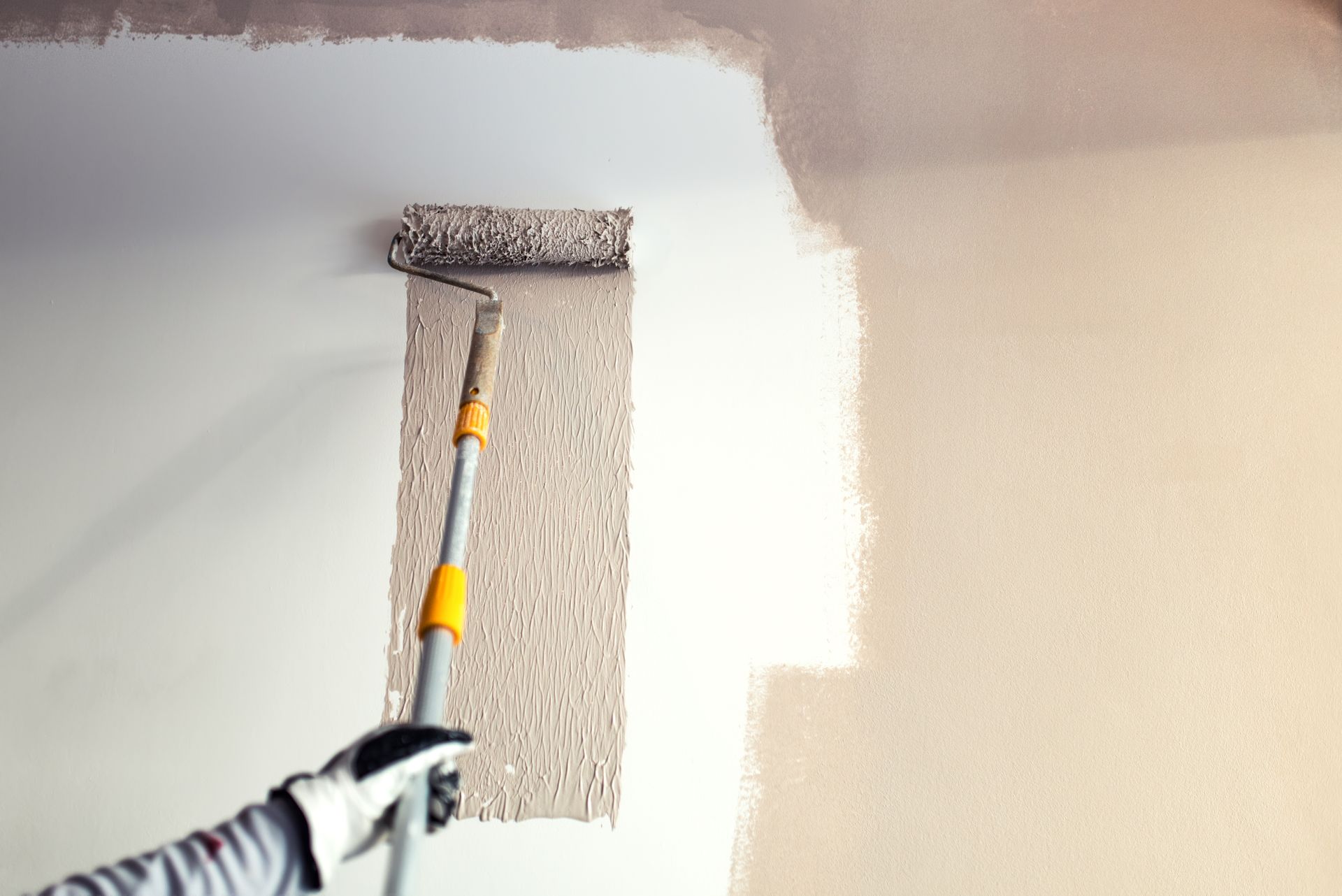 Paint roller applying white paint over a beige wall, partially covering textured wallpaper.