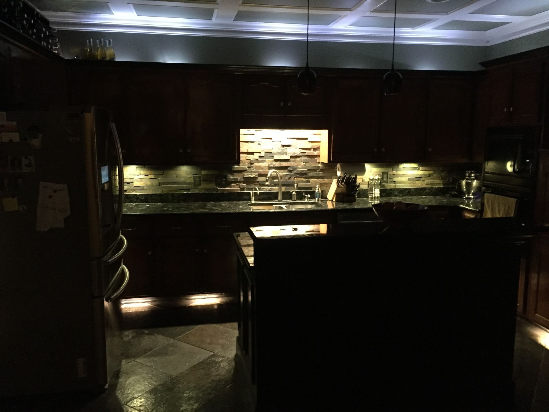 A kitchen with a sink , refrigerator , and microwave is lit up at night.