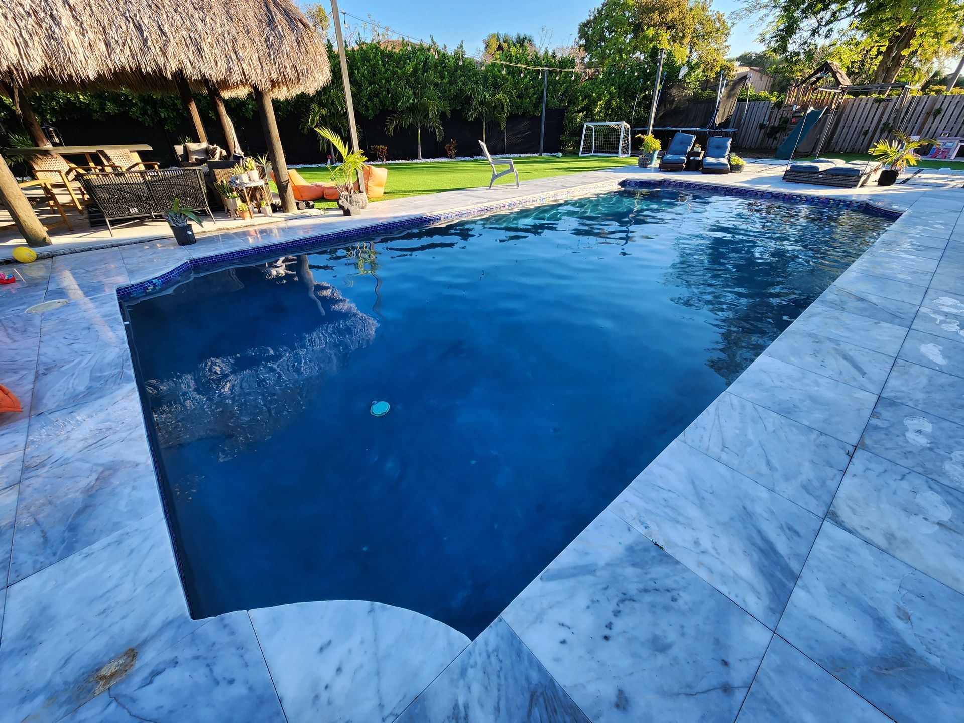 A pool with blue water surrounded by marble tiles, next to a thatch-roofed structure.