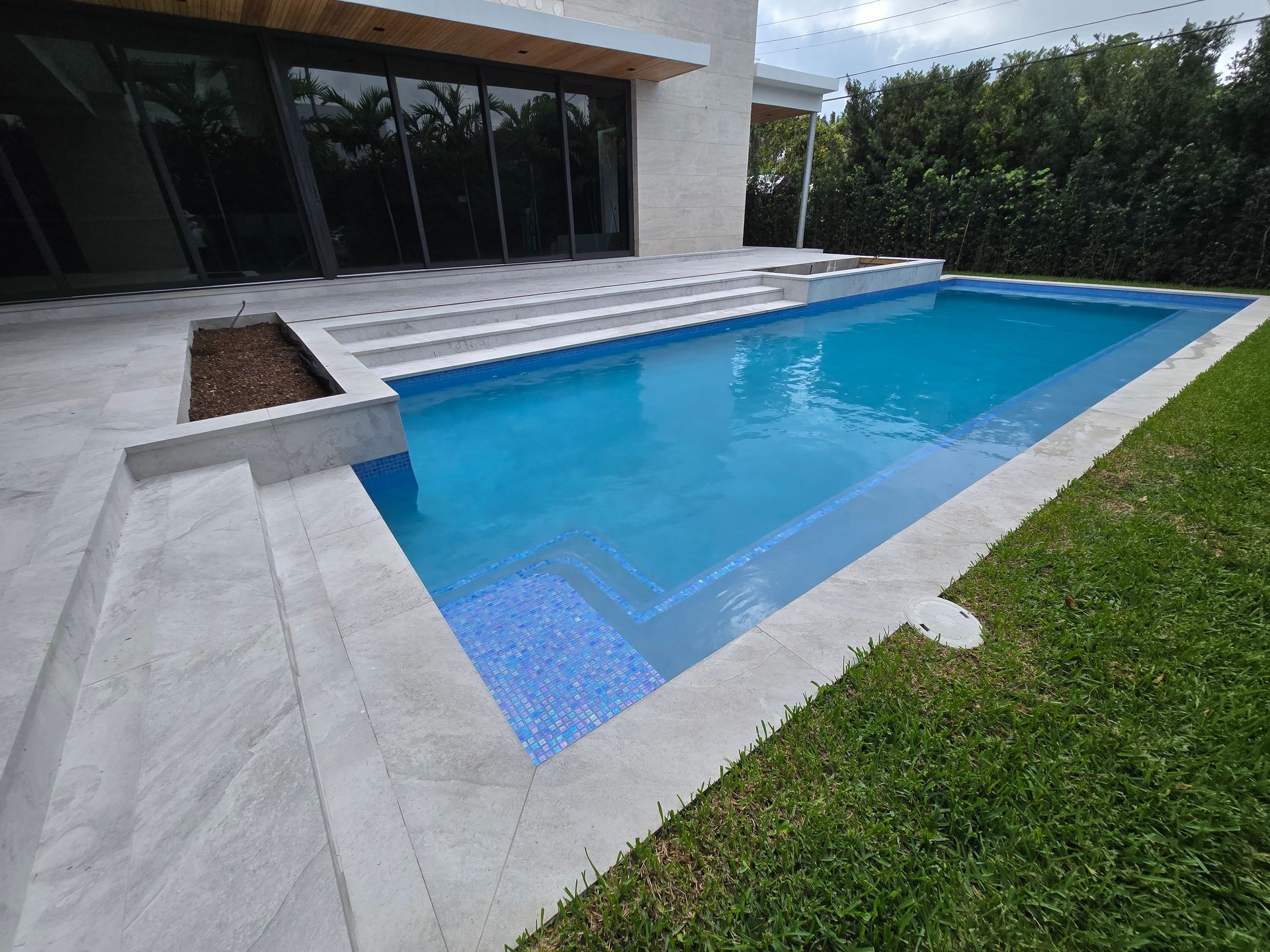 A rectangular pool with blue tile, concrete steps, and green grass next to a modern building.