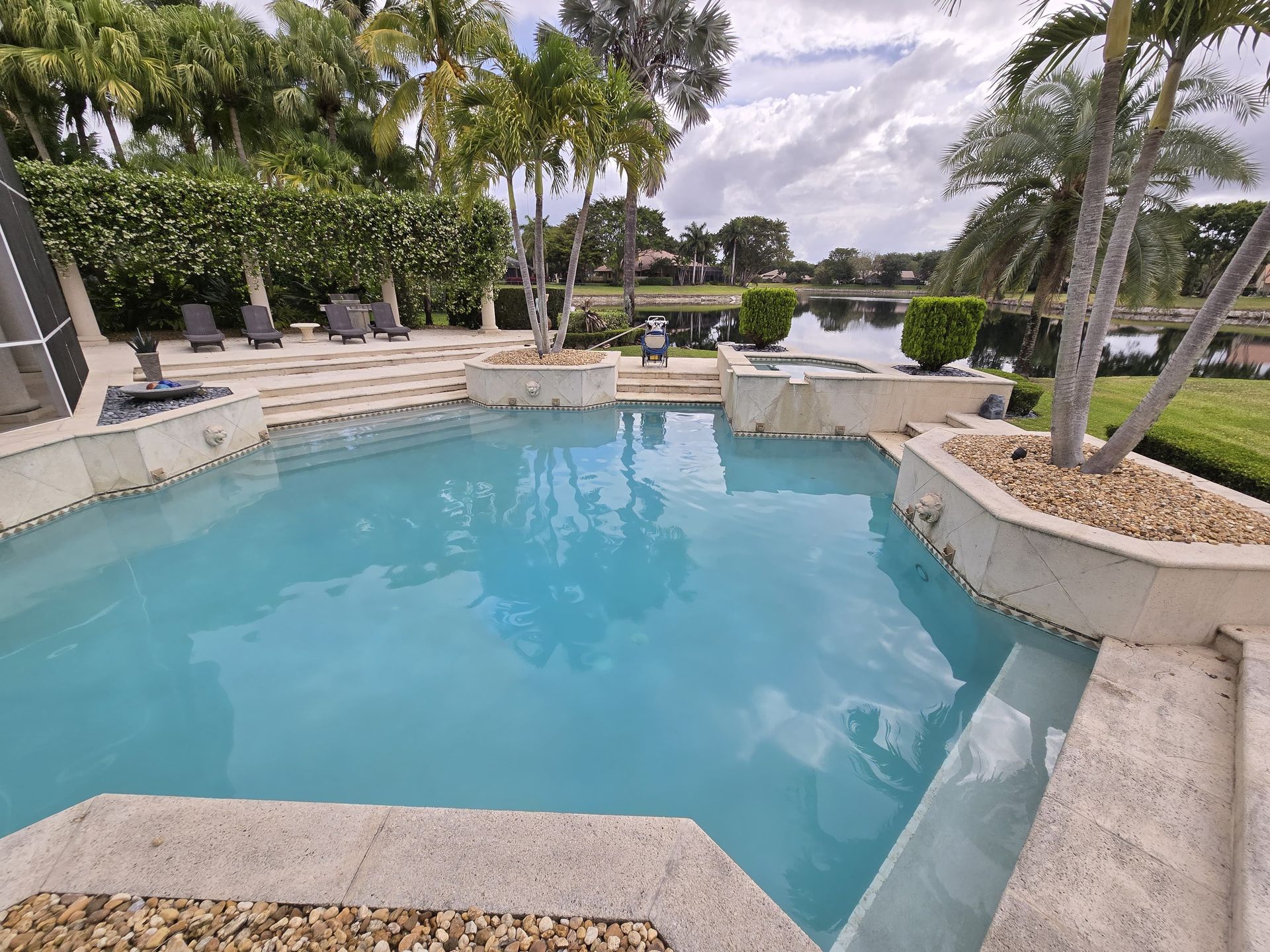 A turquoise swimming pool surrounded by tan stone, overlooking a lake and greenery under a cloudy sky.