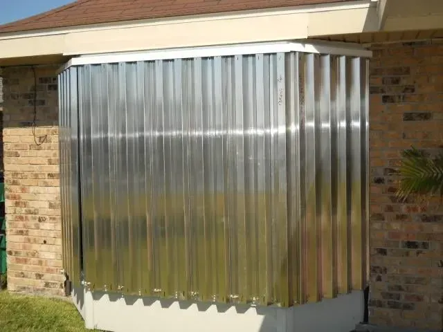 A metal, corrugated shield covers a bay window, protecting it from potential damage; a brick house is in the background.