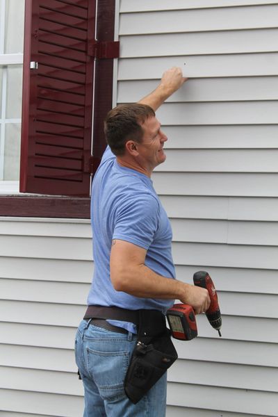 Man in blue shirt and jeans using a power drill on the side of a white house.