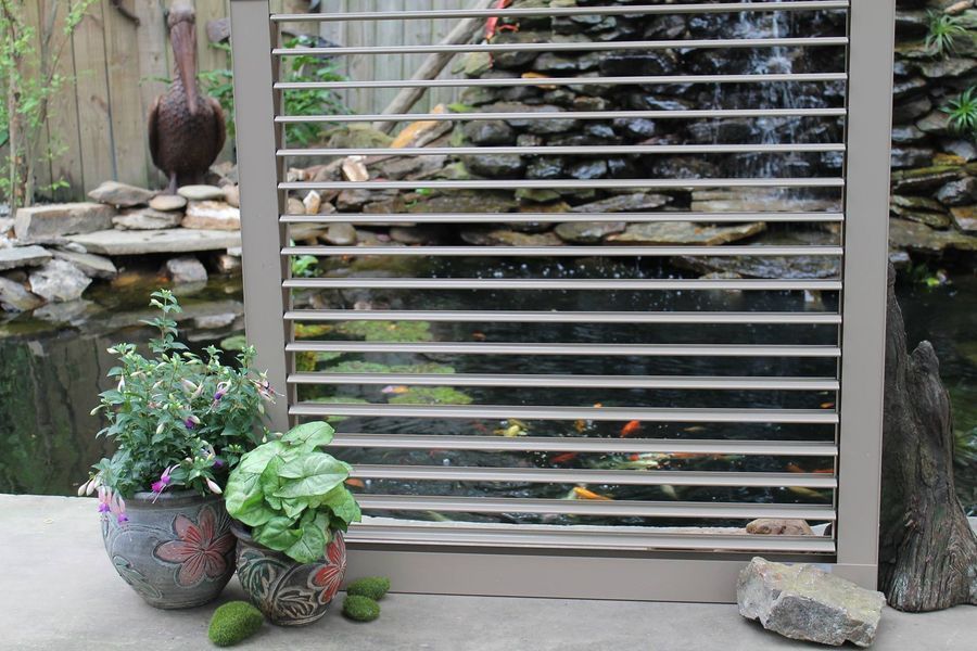 A window with horizontal slats overlooks a pond with a waterfall and plants in decorative pots.