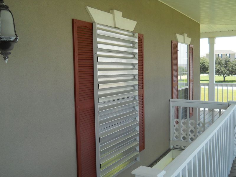 Exterior of house with hurricane shutters over a window, red shutters, and white porch railing.