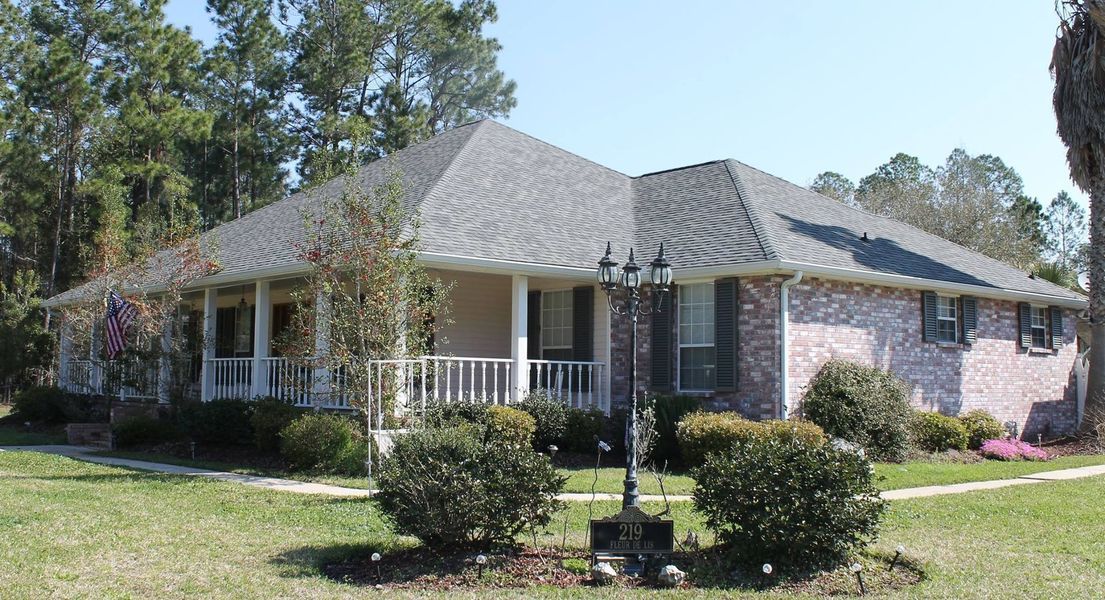 Ranch-style house with porch and brick exterior, surrounded by grass and trees under a blue sky.
