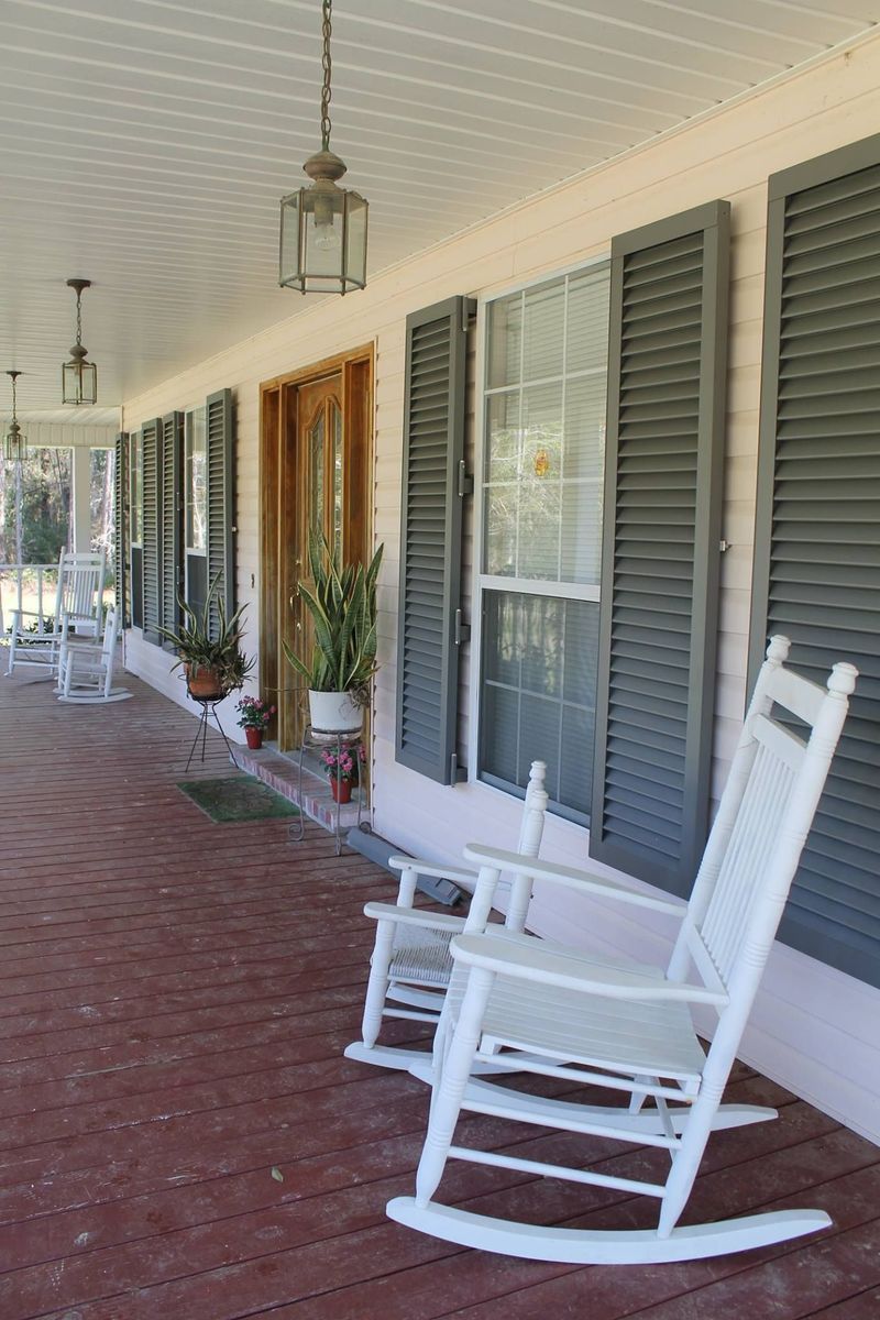 Long porch of a house with white rocking chairs. Wooden door, grey shutters, red floor, and hanging lights.