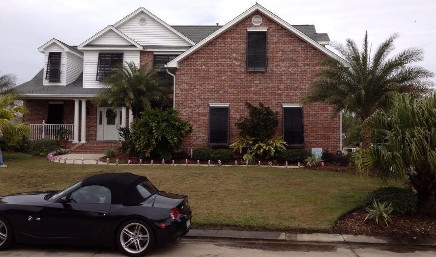 Black convertible parked in front of a two-story brick house with a lawn, palm trees.