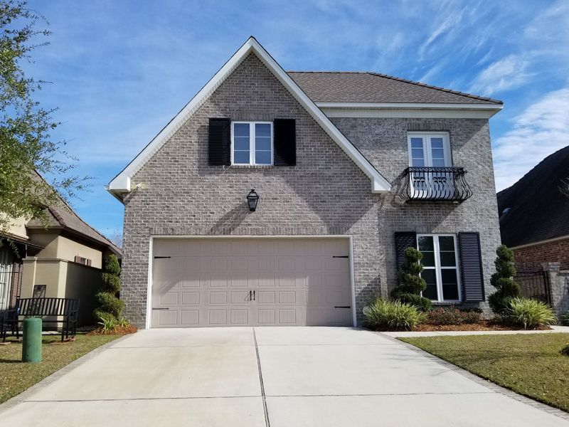 Two-story brick house with beige garage door, black shutters, and small balcony; clear, blue sky.