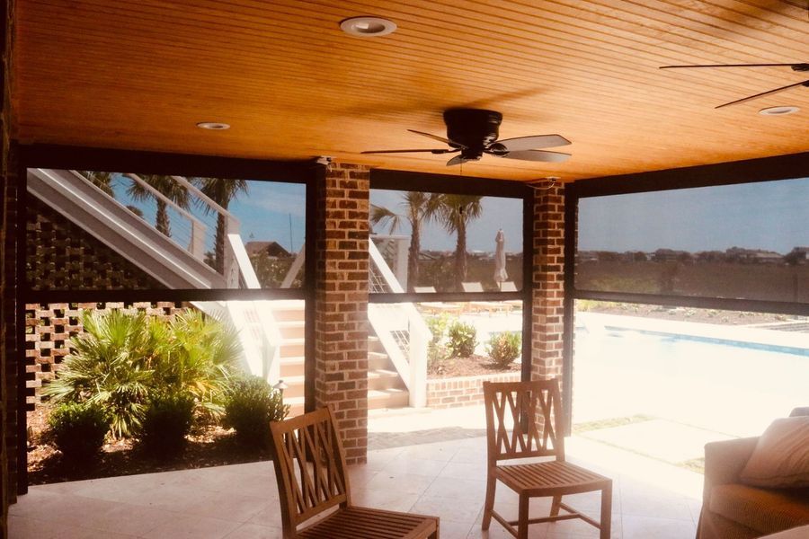 Outdoor patio with brick walls, screened windows, and wood chairs, overlooking a pool and plants.