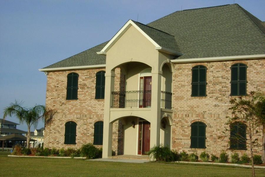 Two-story brick house with green shutters, a balcony, and a green roof on a sunny day.