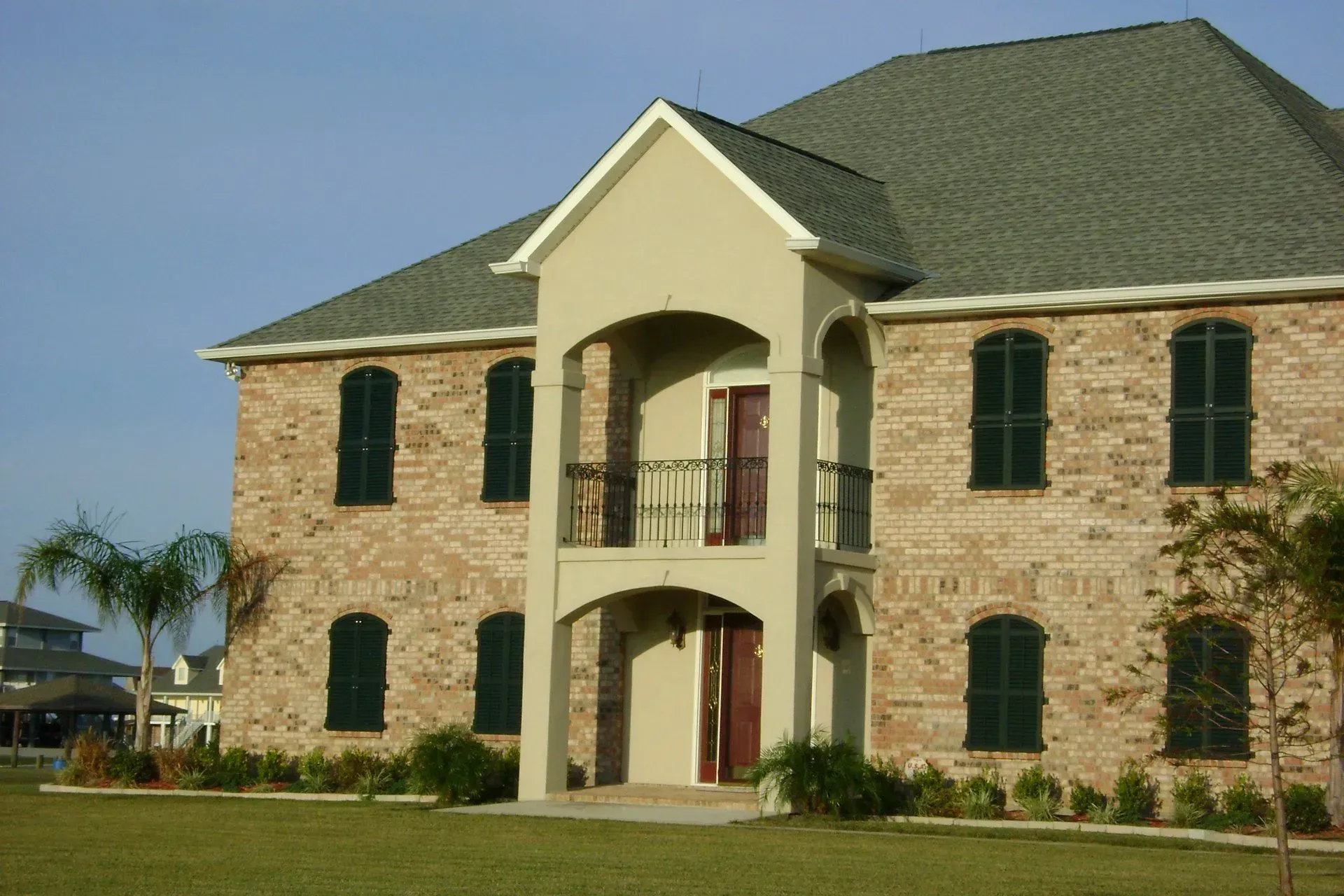 Two-story brick house with green shutters, beige accents, balcony, and green lawn.