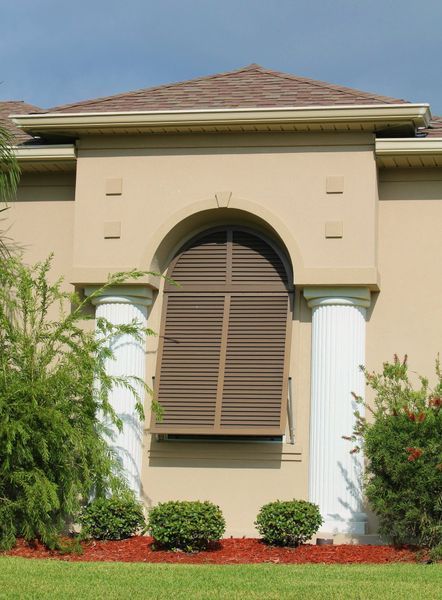 Tan building with arched window and shutters, framed by white pillars and landscaping.