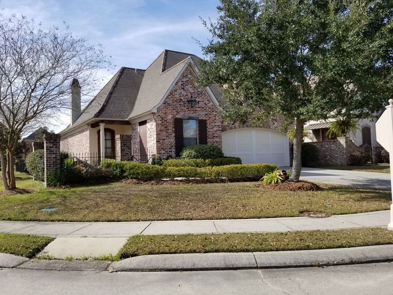Brick house with a brown roof, green bushes, and a driveway, on a sunny day.
