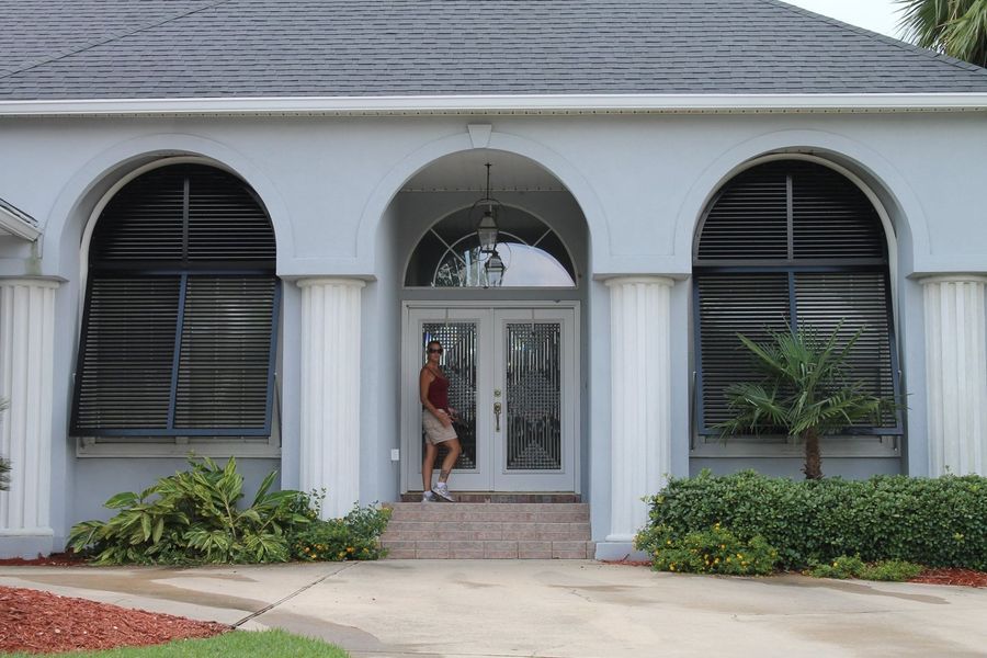 Woman standing in front of a light blue house with arched windows, columns, and a dark roof.