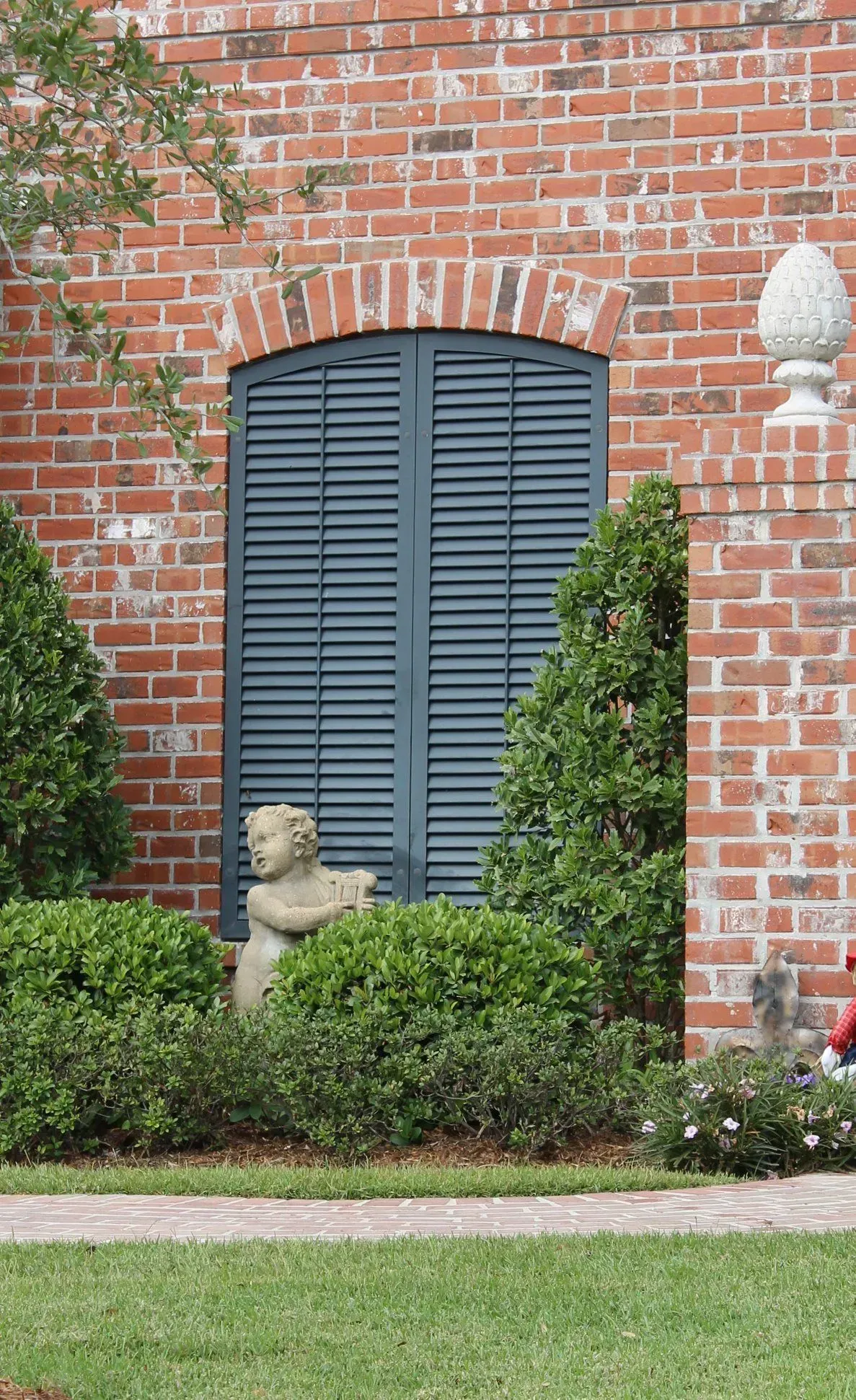 Brick building with arched window covered by blue shutters; statue in garden.