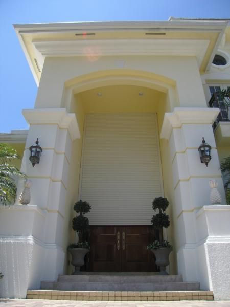 Grand, white entrance with columns, ornate lamps, and double doors; potted topiary flank entrance.