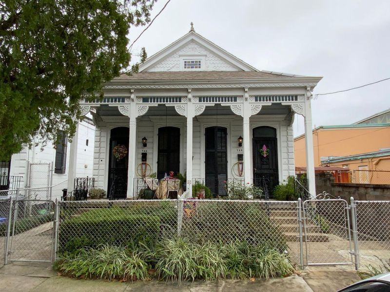 White Creole cottage with porch, black doors, and a chain-link fence, set against a cloudy sky.