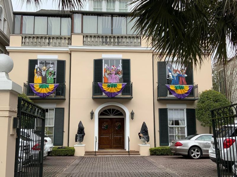 Mardi Gras-decorated beige house with balconies, purple and yellow banners, and three cars parked in the driveway.