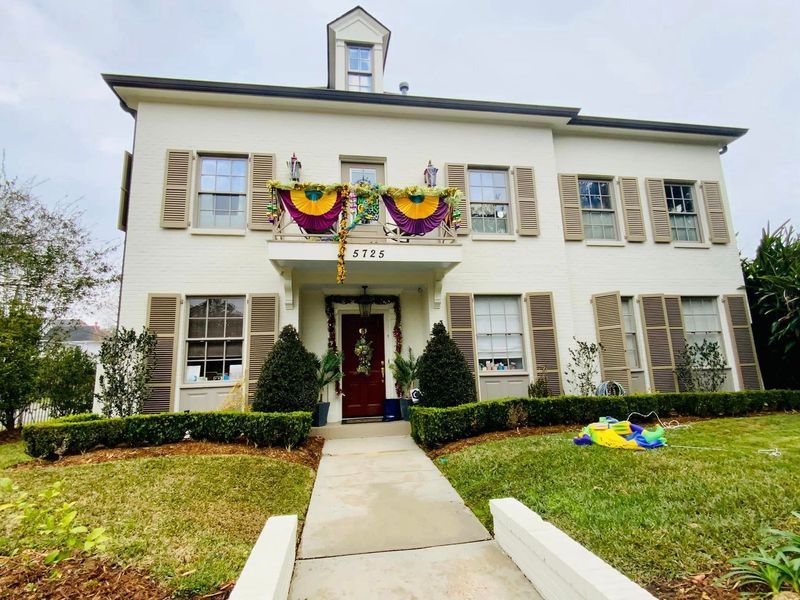 Two-story white house with brown shutters, decorated for Mardi Gras with purple, green, and gold bunting.