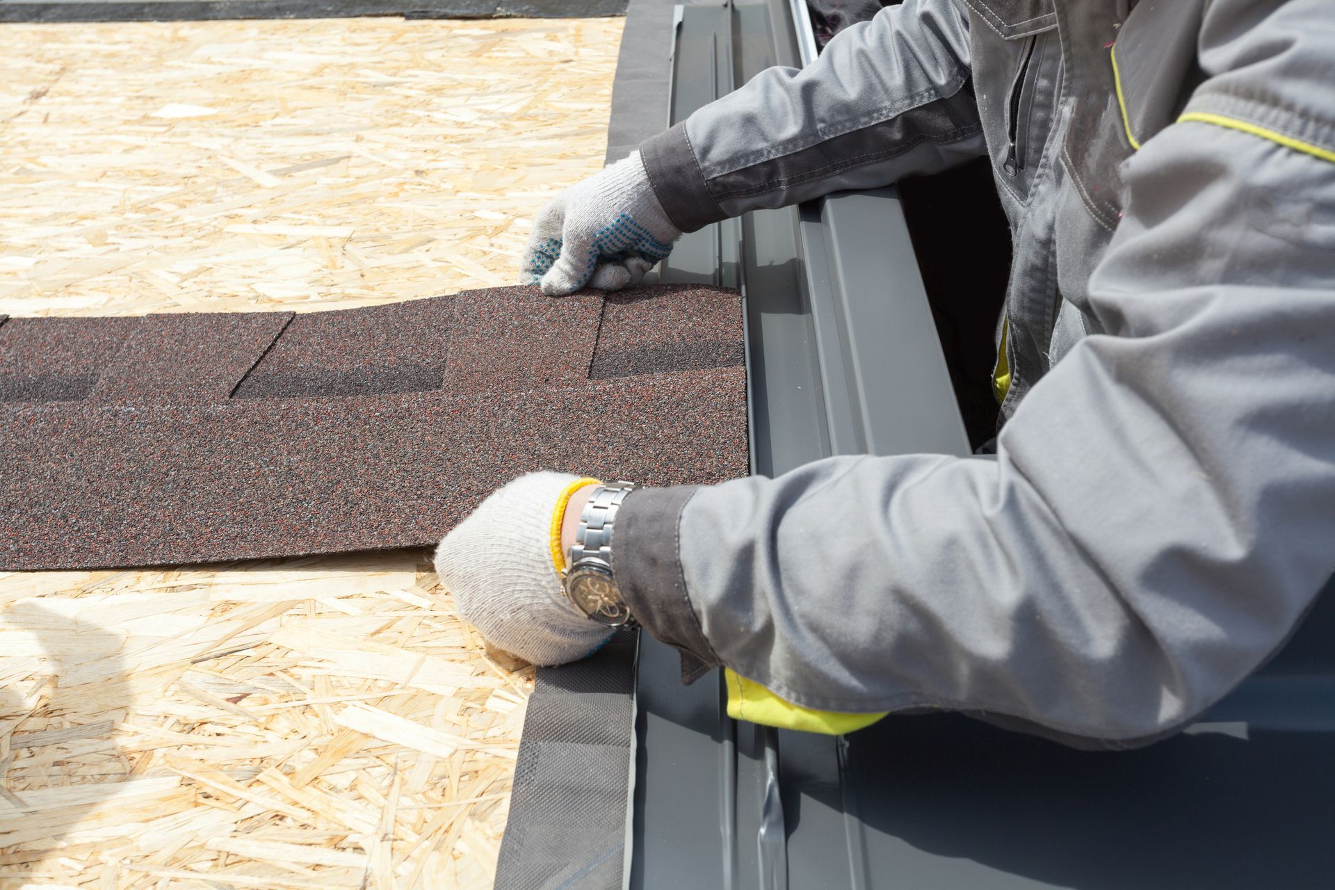 View of a worker wearing a gray uniform and gloves, laying asphalt tile sheet for roof replacement.