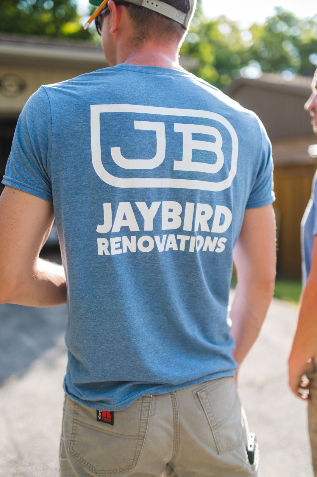 A man wearing a blue jaybird renovations t-shirt is standing in front of a garage.