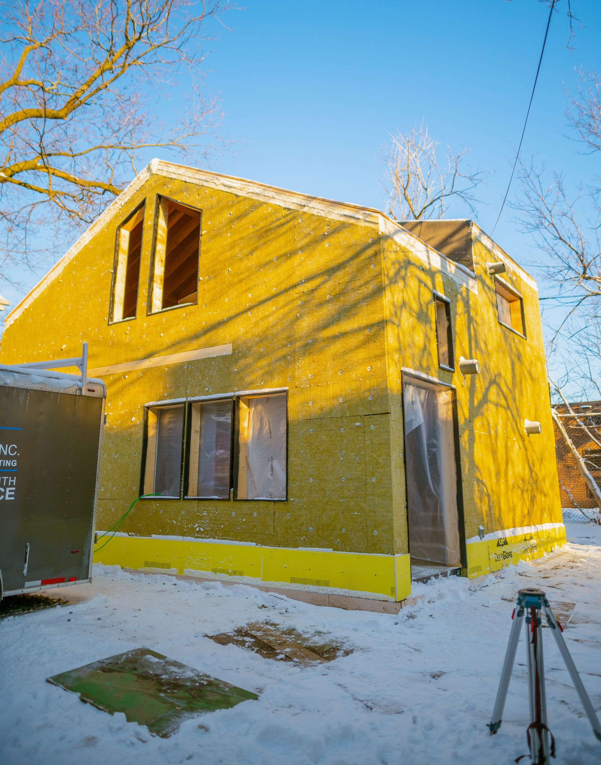 A yellow house is being built in the snow with a tripod in front of it.